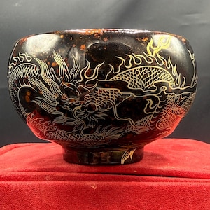 May include: A dark brown bowl with a detailed dragon design in gold and white. The bowl has a rounded shape and sits on a small base. The dragon is intricately carved around the exterior of the bowl.