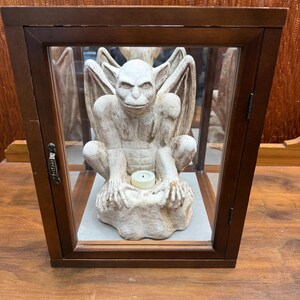 May include: A wooden display case with a glass front houses a stone gargoyle statue. The gargoyle is light-colored, with large wings and a candle on its base. The case has a dark brown frame and a small door.