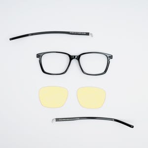 Puede incluir: Gafas negras con lentes transparentes y montura negra. Las gafas están desmontadas, con las patillas y los lentes separados del marco. También se incluyen dos lentes de color amarillo.