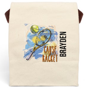 May include: A white canvas tote bag with brown straps. The bag has a tennis racket and ball graphic with the text "CAUSE A RACKET" and the name "BRAYDEN" printed on it.