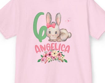 Könnte beinhalten: Ein hellrosa T-Shirt mit einer Grafik eines Hasen mit rosa Schleife, einer großen grünen Zahl sechs, dem Namen ANGELICA in rosa Buchstaben und einem bunten Blumenarrangement am unteren Rand. Das Design ist mittig auf dem T-Shirt platziert.