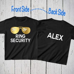 Könnte beinhalten: Ein schwarzes kurzärmeliges T-Shirt, das von vorne und hinten gezeigt wird. Die Vorderseite zeigt eine goldfarbene Sonnenbrille über dem weißen Text RING SECURITY. Die Rückseite zeigt den weißen Text ALEX. Das T-Shirt liegt auf weißem Holz.