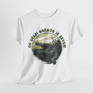 Könnte beinhalten: Ein weißes Kurzarm-T-Shirt mit einer kreisförmigen Grafik eines grünen Alligators, der aus dem Wasser kommt. Der Text "OH SNAP! GREGOR IS SEVEN!" ist in dunkelgrünen Buchstaben am oberen Rand des Designs gedruckt.