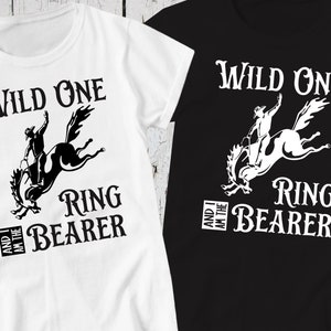 May include: Two white t-shirts with black text and a black silhouette of a cowboy riding a bucking horse. The text on the left shirt reads "Wild One and I am the Ring Bearer". The text on the right shirt reads "Wild One and I am the Ring Bearer".