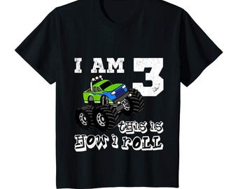 Könnte beinhalten: Ein schwarzes Kurzarm-T-Shirt mit einer Grafik eines grünen und blauen Monstertrucks. Der Text lautet I AM 3 und THIS IS HOW I ROLL in weißer, abgenutzter Schrift. Das Design enthält eine große Zahl 3 und einen Cartoon-Truck mit großen Reifen.