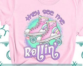 May include: A light pink t-shirt featuring a graphic of two pink roller skates with rainbow accents, clouds, and hearts. The text "they see me" is printed in purple above the skates, and "Rollin'" is printed in large purple letters below.