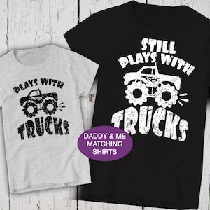 May include: A light gray t-shirt with black text reading PLAYS WITH TRUCKS and a monster truck graphic, alongside a black t-shirt with white text reading STILL PLAYS WITH TRUCKS and a monster truck graphic, featuring a purple DADDY & ME MATCHING SHIRTS label.