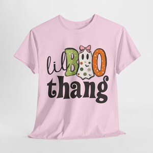 May include: A light pink short-sleeve t-shirt featuring a graphic print with the text 'lil BOO thang'. The design includes a green letter B, an orange letter O, and a white ghost character wearing a pink bow, decorated with small flowers and bats.