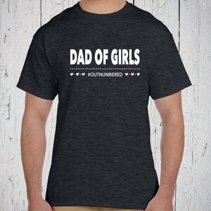 Könnte beinhalten: Ein dunkelgrau meliertes Kurzarm-T-Shirt mit dem weißen Schriftzug DAD OF GIRLS in fetter serifenloser Schrift, gefolgt von einer gepunkteten Linie und dem Text #OUTNUMBERED, flankiert von kleinen weißen Herzsymbolen.