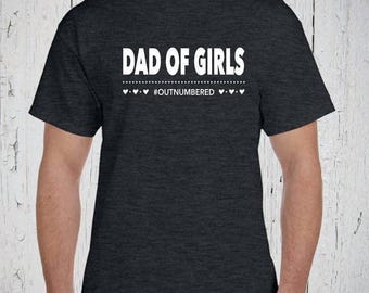 Könnte beinhalten: Ein dunkelgrau meliertes Kurzarm-T-Shirt mit dem weißen Schriftzug DAD OF GIRLS in fetter serifenloser Schrift, gefolgt von einer gepunkteten Linie und dem Text #OUTNUMBERED, flankiert von kleinen weißen Herzsymbolen.