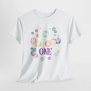 May include: A white short-sleeved t-shirt featuring a graphic design of colorful watercolor bubbles. The text "the BUBBLY ONE" is printed in the center, with the letters B, U, B, L, Y, and ONE integrated into the bubble pattern in purple and white.