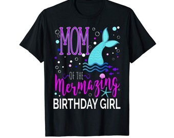 May include: A black short-sleeve t-shirt featuring a graphic print with the text MOM OF THE Mermazing BIRTHDAY GIRL. The design includes a light blue mermaid tail, purple lettering, white bubbles, a pink seashell, and a light blue starfish.