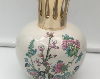 Könnte beinhalten: Eine cremefarbene, runde Öllampe aus Keramik mit einem goldfarbenen Metallaufsatz. Die Lampe ist mit einem Blumendesign in Rosa, Blau und Grün verziert. Das Design zeigt Blumen und Zweige.
