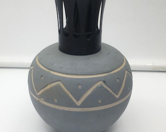 Könnte beinhalten: Eine graue Keramik-Aromatherapie-Lampe mit schwarzem Deckel. Die Lampe hat einen runden Sockel mit einem geometrischen Design in einer helleren Farbe. Der Deckel hat eine Reihe vertikaler Schlitze.