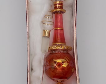 Puede incluir: Un frasco de perfume de vidrio rojo con detalles dorados y un gotero de vidrio transparente en una caja roja y blanca. El frasco tiene un cuerpo redondo y un tapón dorado.