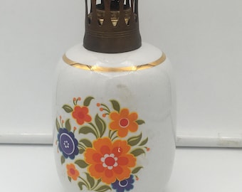 Könnte beinhalten: Eine Vintage-Öllampe aus weißer Keramik mit einem Blumendesign in Orange, Rot und Blau. Die Lampe hat einen goldenen Rand und einen bronzefarbenen Metallaufsatz. Die Lampe ist ein dekorativer Gegenstand.