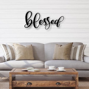 May include: A black metal wall decor with the word "blessed" in a cursive font. The decor is hanging on a white shiplap wall above a gray couch with pillows.