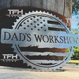 May include: A metal sign with a distressed American flag design and the text "DAD'S WORKSHOP" in a circular shape with a gear-like border.