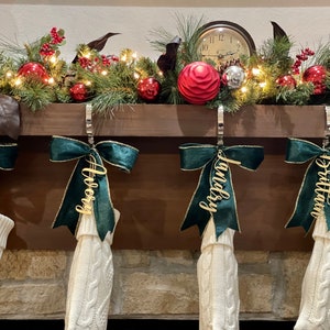 May include: Four white knitted stockings with green velvet bows and gold personalized name tags hanging on a fireplace mantel. The names on the tags are "Lauren", "Avery", "Landry", and "Brittan".