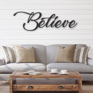 May include: A gray couch with pillows sits in front of a white wood wall with the word "Believe" in black cursive script.