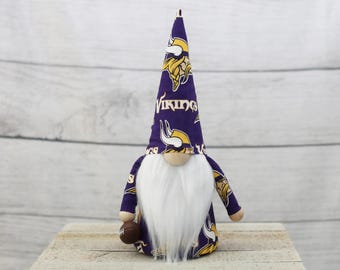 May include: A decorative gnome doll featuring a long white beard, a tan nose, and a tall purple hat and matching outfit. The hat and outfit are patterned with the Minnesota Vikings logo and the word "Vikings". The gnome is holding a football.