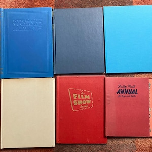 Puede incluir: Una colección de libros antiguos con tapas duras en varios colores. Los libros incluyen un atlas azul, un libro azul marino, un libro azul claro, un libro color crema, un "Film Show Annual" rojo y un "Daily Mail Annual" rojo.