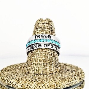 May include: Two silver rings with the name "Tessa" and "Class of 2022" engraved on them. The ring with the name "Tessa" has a row of light blue gemstones.