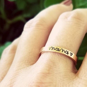 May include: A gold-colored ring with the word "mama" inscribed in a handwritten font, followed by a small heart symbol. The ring is worn on a finger, showcasing its delicate design and sentimental message.