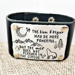 Pode incluir: Pulseira de couro preto com uma placa de metal prateada gravada com a citação "O leão e o tigre podem ser mais poderosos... Mas o lobo não se apresenta no circo". A placa também apresenta uma lua crescente, montanhas, árvores e um lobo.