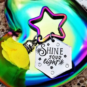 May include: A silver keychain with a star charm and a hexagonal charm that says "Shine your light". The keychain is on a rainbow iridescent heart-shaped dish.