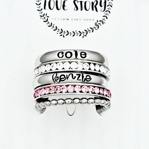 May include: A set of four silver rings with the names "Cole" and "Kenzie" engraved on them. The rings are decorated with clear and pink crystals.