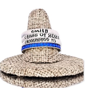 May include: A silver ring set with three bands. The top band says "emily", the middle band says "class of 2023", and the bottom band says "Okeechobee HS". The bottom band is accented with blue rhinestones.