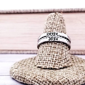 May include: A silver ring engraved with the names "RAVEN" and "JOSH". The ring features a central braided pattern. The ring is presented on a beige ring display.