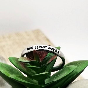 Op de afbeelding: Zilveren ring met de woorden "use your words" gegraveerd erop. De ring ligt op een groene succulent.