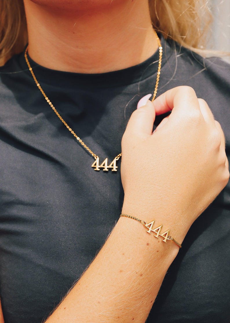 Op de afbeelding: Goudkleurige ketting- en armbandset. De ketting heeft het nummer "444" als hanger en de armband toont ook "444". Beide stukken zitten aan een delicate ketting, waardoor een minimalistische sieradenset ontstaat.