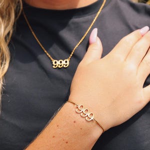 Op de afbeelding: Goudkleurige ketting- en armbandset. De ketting heeft het nummer "999" als hanger en de armband toont ook "999". Beide stukken zijn aan fijne kettingen, wat een minimalistische en elegante uitstraling cre&euml;ert. De sieraden staan tegen een donkere achtergrond.