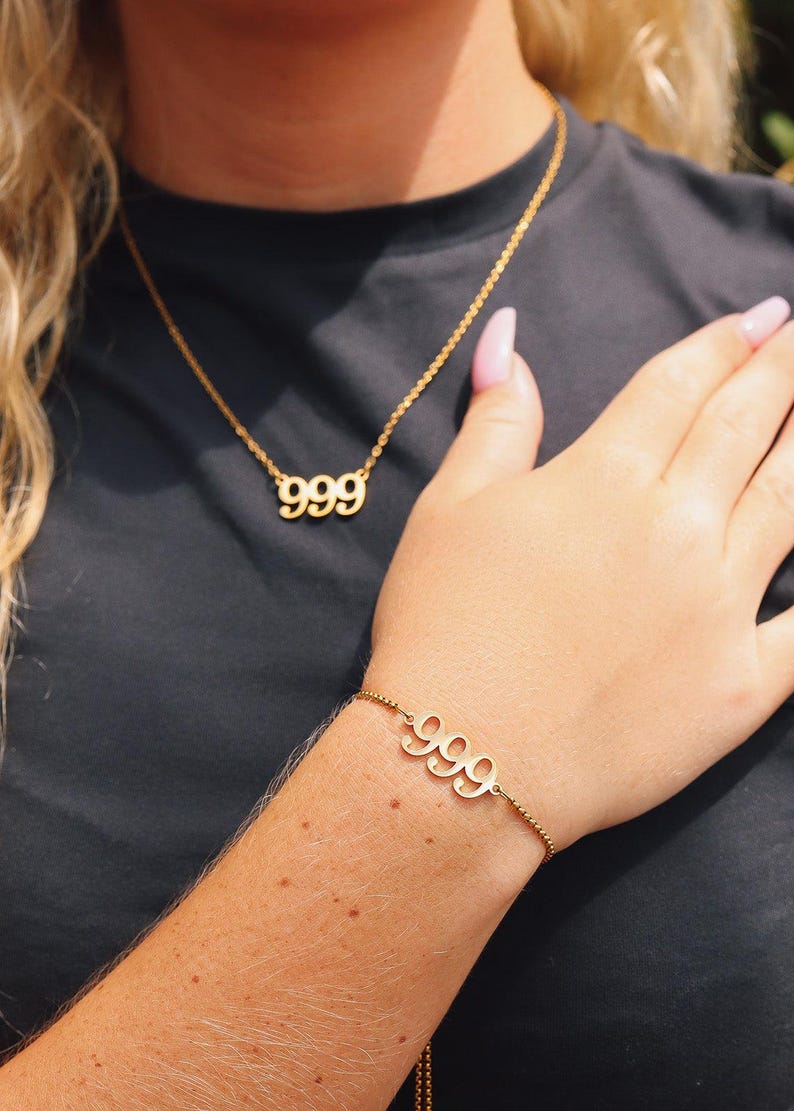 Op de afbeelding: Goudkleurige ketting- en armbandset. De ketting heeft het nummer "999" als hanger en de armband toont ook "999". Beide stukken zijn aan fijne kettingen, wat een minimalistische en elegante uitstraling cre&euml;ert. De sieraden staan tegen een donkere achtergrond.