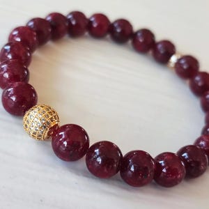 Op de afbeelding: Een armband met grote, ronde, dieprode kralen. Een goudkleurige kraal, bezet met kleine, heldere steentjes, zorgt voor een accent. De armband wordt gepresenteerd op een lichtgekleurd oppervlak.