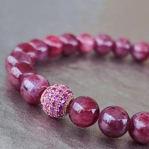 Könnte beinhalten: Nahaufnahme eines Armbands mit tiefroten, runden Edelsteinen. Eine einzelne, größere Perle ist mit kleineren, funkelnden rosa Steinen besetzt. Das Armband wird auf einer neutralen, strukturierten Oberfläche präsentiert, die die Farben und das Design hervorhebt.