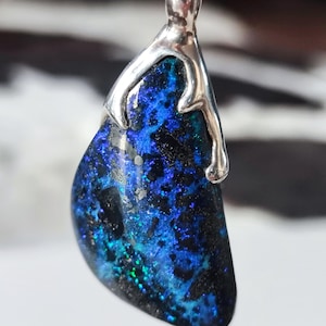 Può includere: Un ciondolo con una grande pietra preziosa irregolare, blu e nera. La pietra ha una qualità scintillante e iridescente con macchie verdi e argentate. È incastonata in una montatura in metallo argentato e pende da una catena di perline.