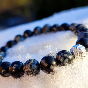 Könnte beinhalten: Armband mit schwarzen und grauen Schneeflocken-Obsidian-Perlen. Das Armband hat eine dekorative Silberperle mit detailliertem Design. Das Armband wird auf Schnee gezeigt. Die Perlen haben einen Durchmesser von etwa 1 cm.