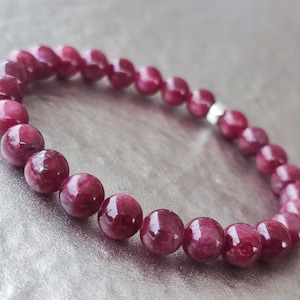 May include: A bracelet made of deep red, polished gemstones. The round beads are strung together with a small silver-coloured spacer bead. The gemstones have a glossy finish. The bracelet is approximately 19 cm in circumference.