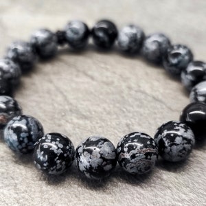 Könnte beinhalten: Ein schwarzes und weißes Schneeflocken-Obsidian-Armband mit runden Perlen. Das Armband besteht aus natürlichen Steinen und ist etwa 20 cm lang.