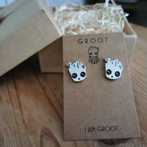 Op de afbeelding: Zilverkleurige Groot-oorstekers op een bruine kaart met de tekst "GROOT" en "I AM GROOT". De oorbellen hebben de vorm van het personage Groot uit Guardians of the Galaxy. De oorbellen worden gepresenteerd in een bruine geschenkdoos.