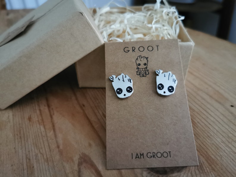 Op de afbeelding: Zilverkleurige Groot-oorstekers op een bruine kaart met de tekst "GROOT" en "I AM GROOT". De oorbellen hebben de vorm van het personage Groot uit Guardians of the Galaxy. De oorbellen worden gepresenteerd in een bruine geschenkdoos.