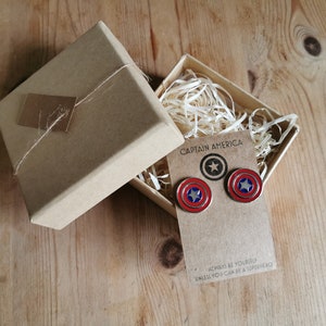 Op de afbeelding: Een Captain America thema cadeau set. De set bevat een paar schild oorbellen en een bijpassende pin, gepresenteerd in een kraft papieren doos met decoratieve vulling. De oorbellen en pin hebben rode, witte en blauwe kleuren.