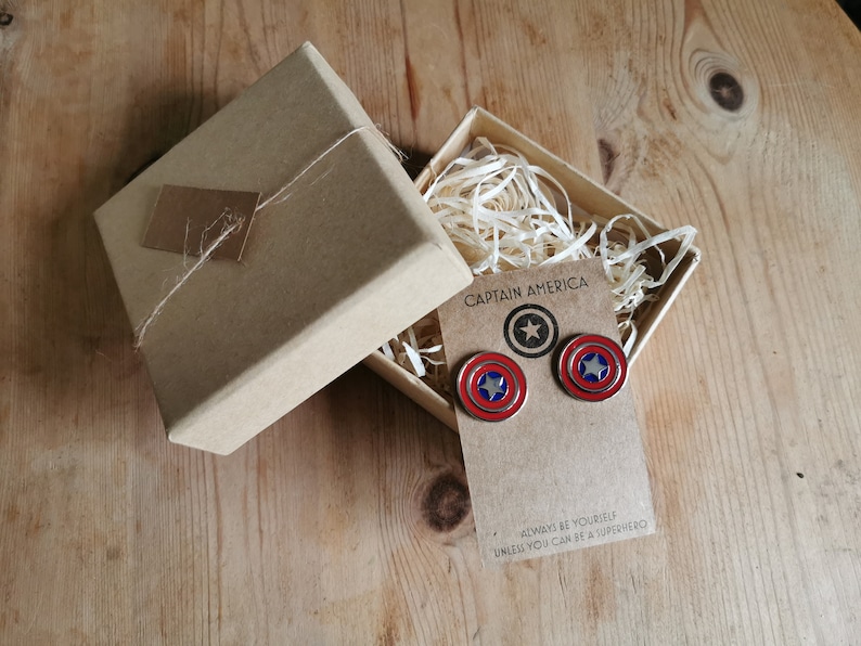 Op de afbeelding: Een Captain America thema cadeau set. De set bevat een paar schild oorbellen en een bijpassende pin, gepresenteerd in een kraft papieren doos met decoratieve vulling. De oorbellen en pin hebben rode, witte en blauwe kleuren.