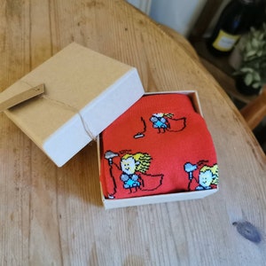 May include: A red gift box with a white lid, containing a pair of socks with a cartoon design of people holding microphones. The socks are red with white and yellow details.