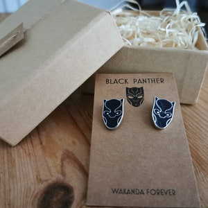 Puede incluir: Un par de pendientes de Black Panther negros y plateados con el texto "Black Panther" y "Wakanda Forever" en una tarjeta marr&oacute;n.