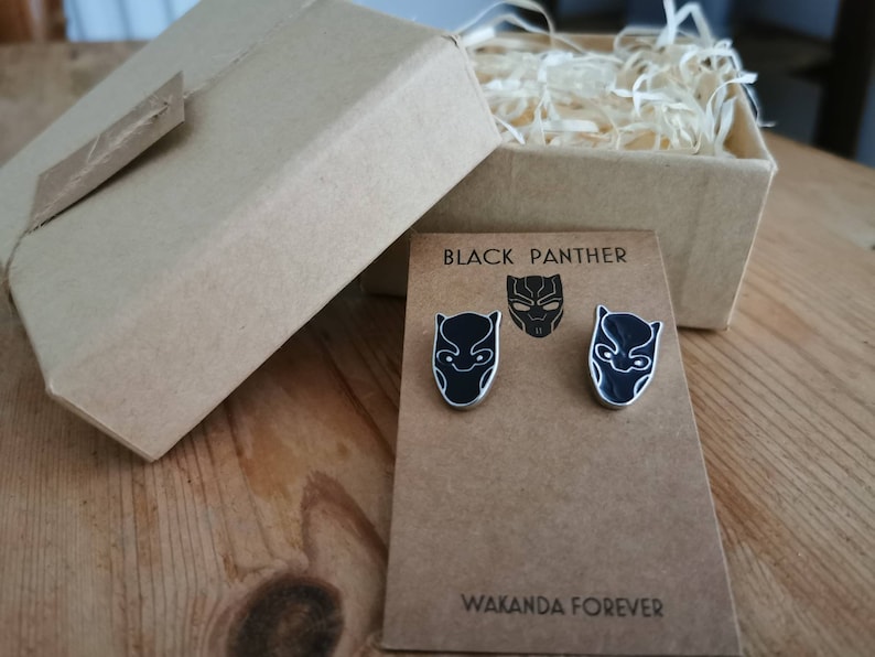 Puede incluir: Un par de pendientes de Black Panther negros y plateados con el texto "Black Panther" y "Wakanda Forever" en una tarjeta marr&oacute;n.